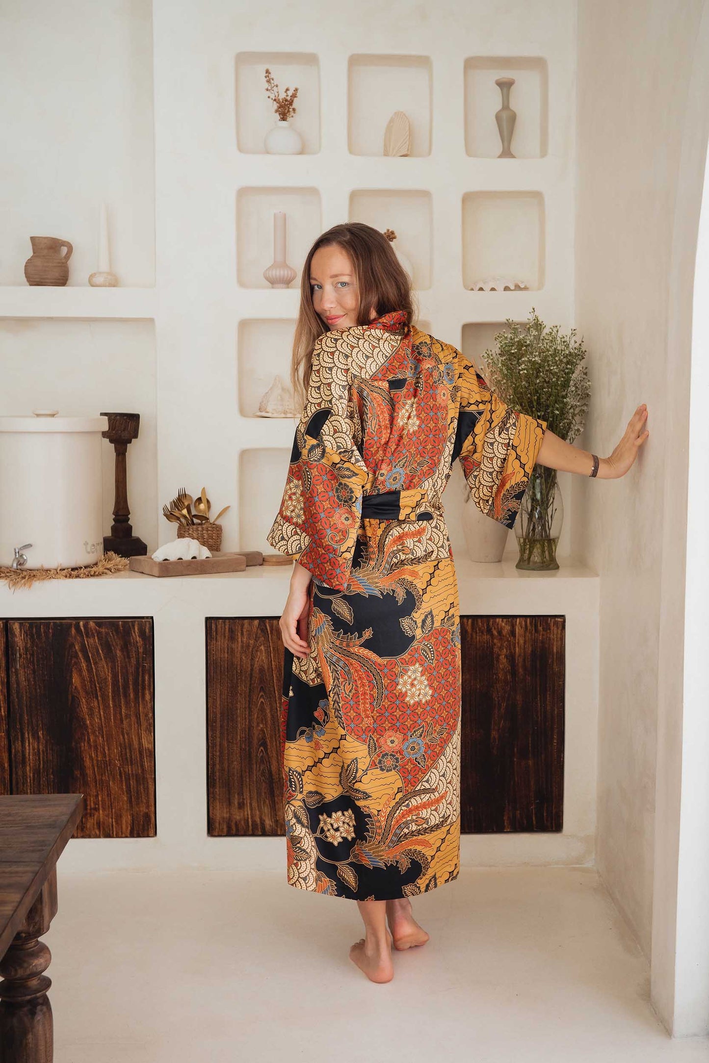 Woman wearing the Agni women’s kimono robe – long satin dressing gown in orange and gold batik design, handmade in Bali.