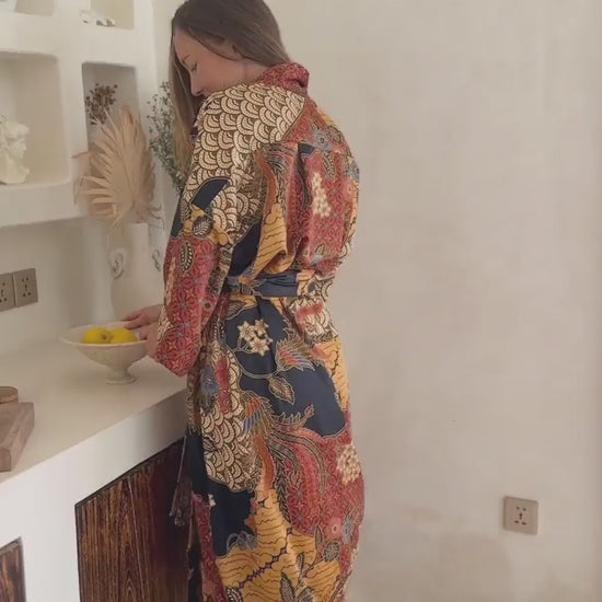 a video of a Woman wearing the Agni women’s kimono robe – long satin dressing gown in orange and gold batik design, handmade in Bali.