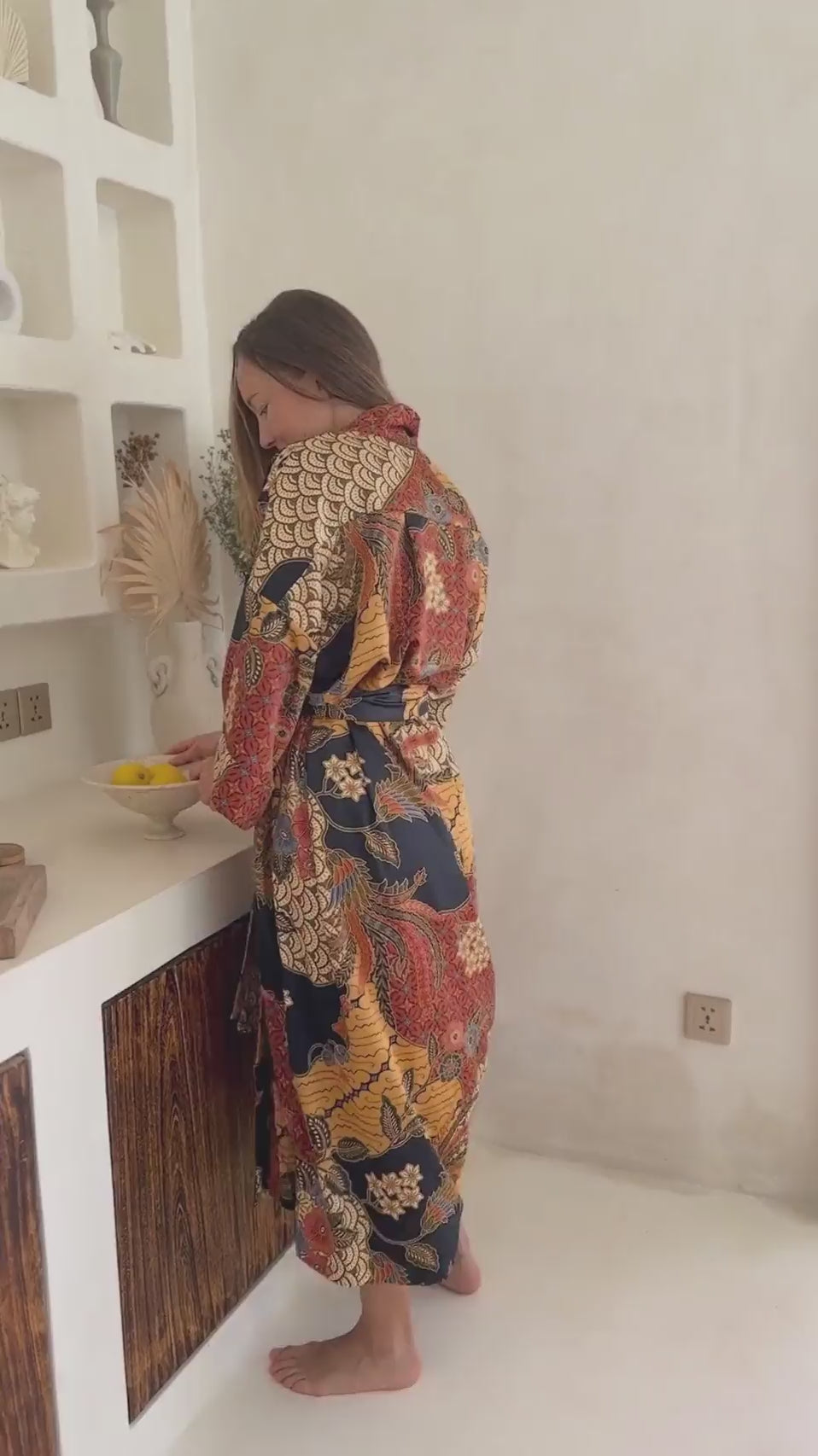a video of a Woman wearing the Agni women’s kimono robe – long satin dressing gown in orange and gold batik design, handmade in Bali.