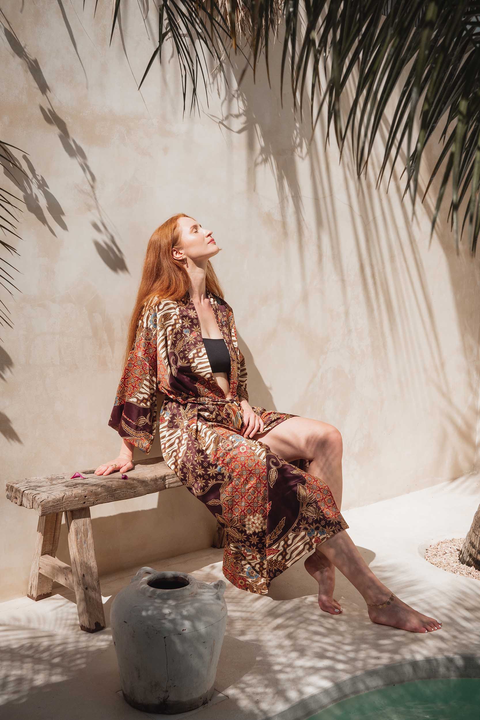 Woman wearing Toré Ratna kimono robe – long burgundy satin dressing gown with golden and cream batik design.