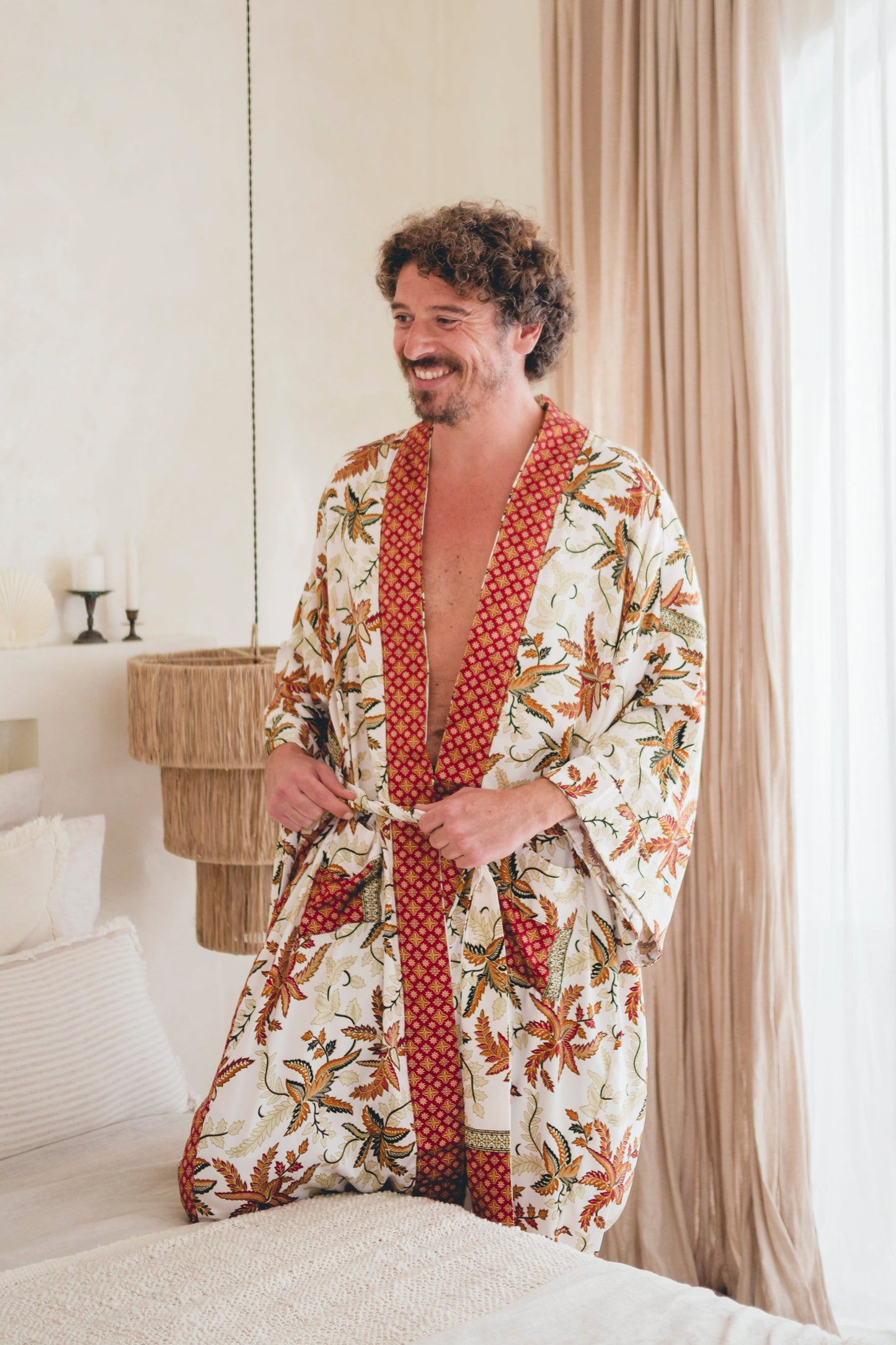 A man wearing a white and red batik patterned kimono robe with red and green botanical motifs and a red patterned trim, standing on a bed in a softly lit bedroom.