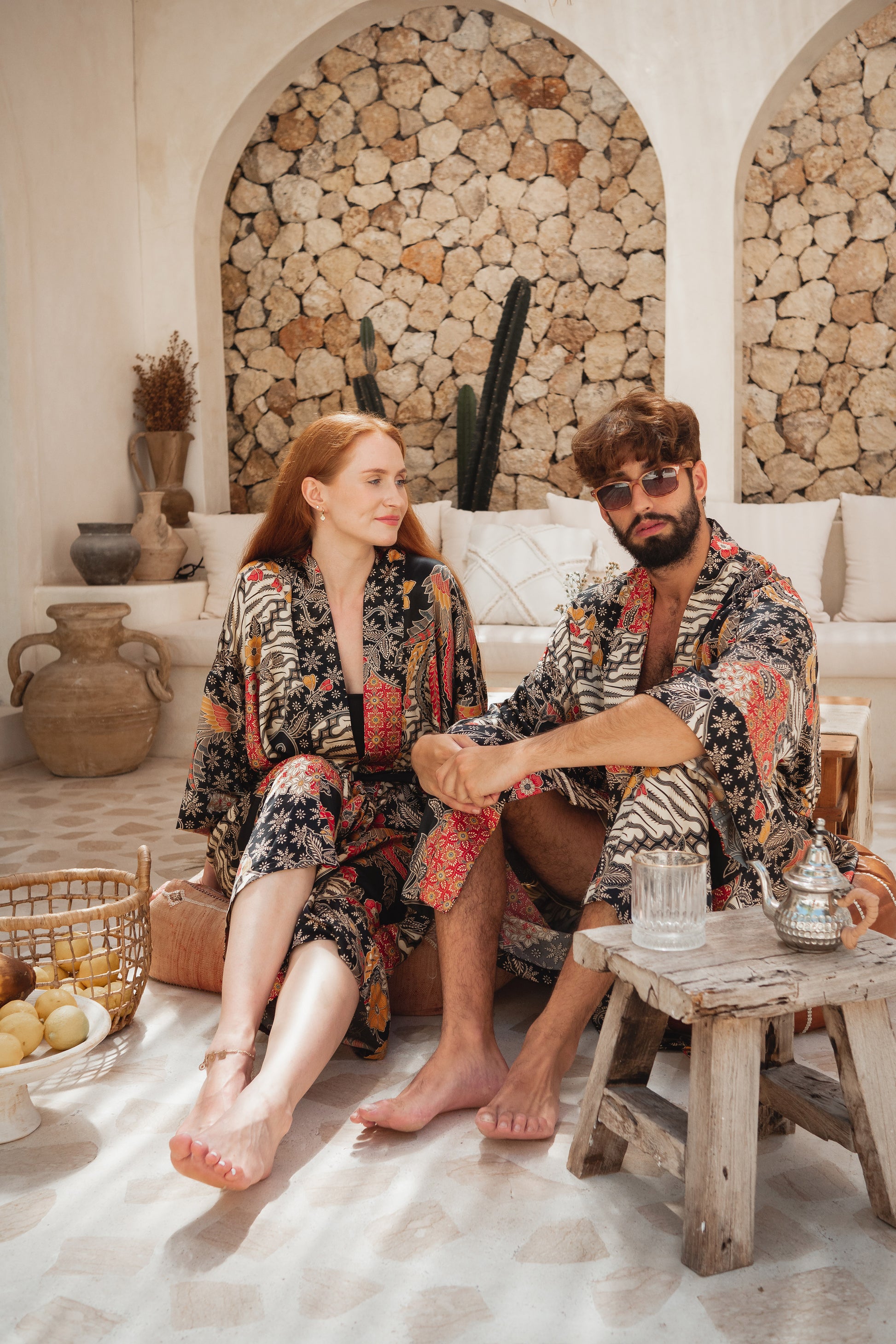 A man and a woman sitting together in a rustic, sunlit stone-walled room, wearing matching patterned bathrobes with black, red, and beige motifs. A small wooden table with glasses and a metal teapot sits in the foreground.