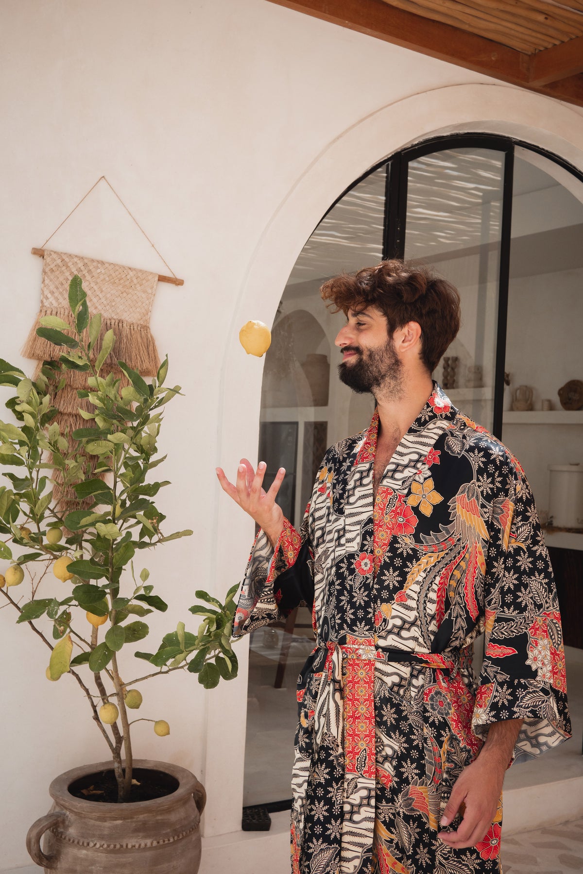 A man wearing a black and white patterned kimono robe with red, orange, and yellow floral accents, standing in a bright room with a potted lemon tree and an arched doorway.