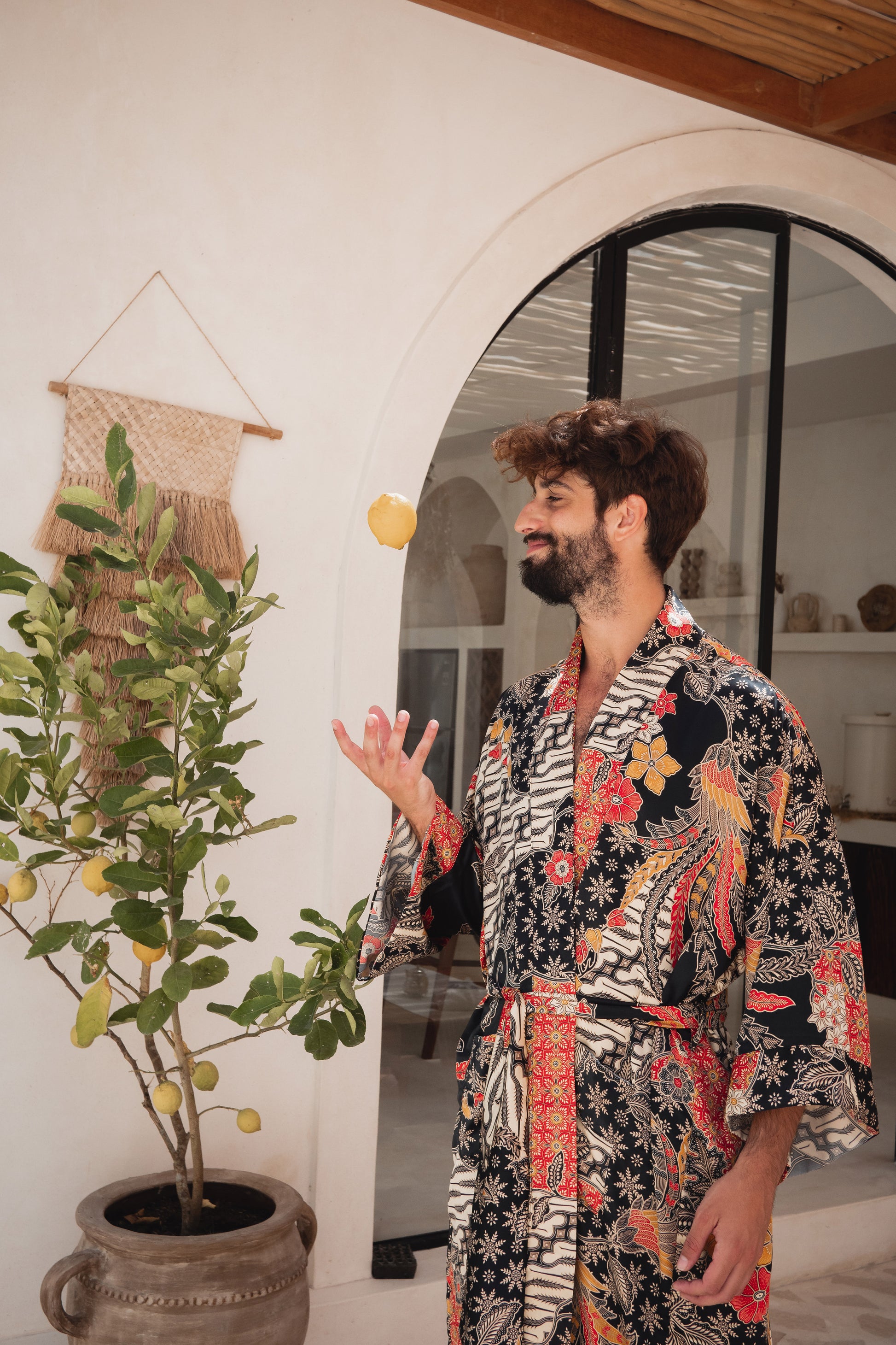 A man wearing a black and white patterned kimono robe with red, orange, and yellow floral accents, standing in a bright room with a potted lemon tree and an arched doorway.