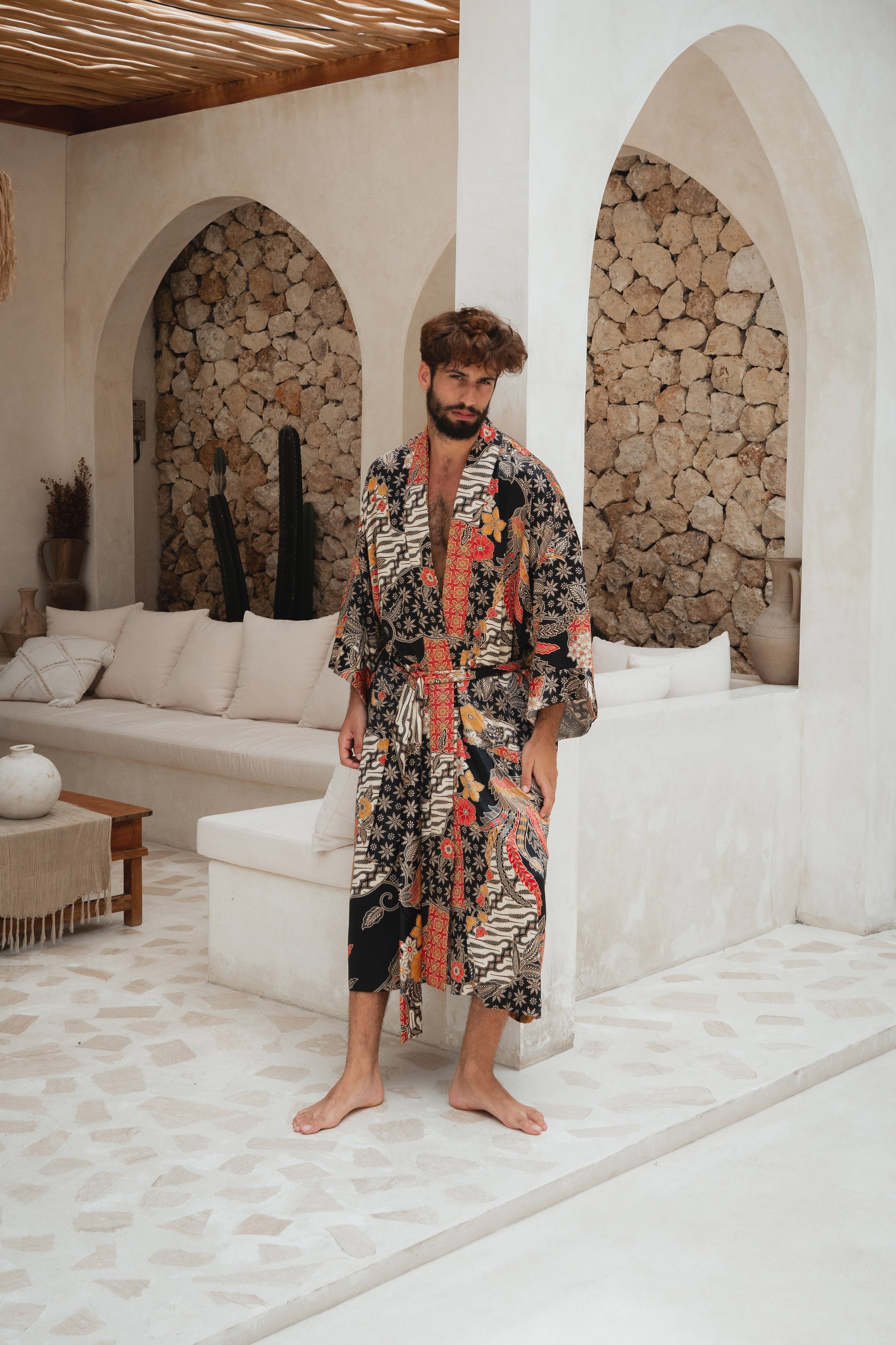 A man wearing a long-sleeve patterned robe stands barefoot in a bright, modern outdoor lounge area with white walls, stone arches, and a sofa in the background.