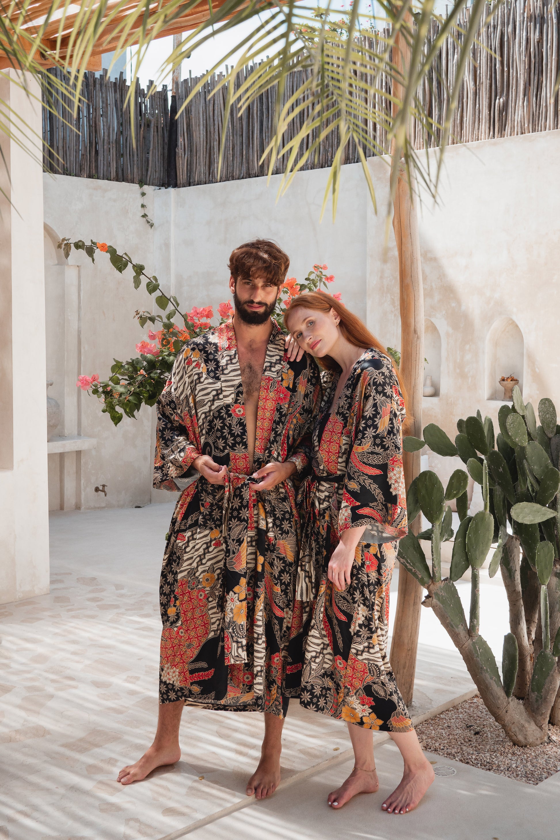 A man and a woman wearing matching tropical print bathrobes stand barefoot in a sunlit outdoor patio setting with palm leaves, cactus plants, and white stucco walls.