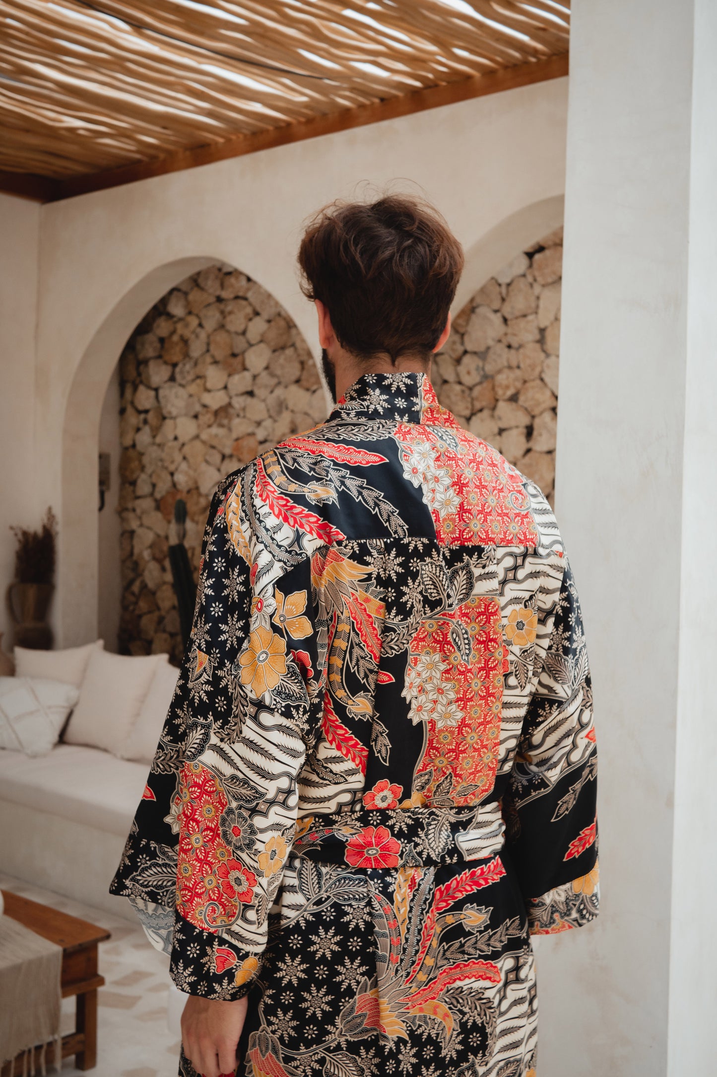 A man viewed from behind wearing a long-sleeve patterned robe with a black base and colorful floral, and red-orange motifs, standing in a rustic indoor setting with stone arches and a woven ceiling.