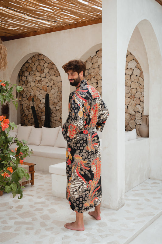 A man wearing a long-sleeve patterned robe stands barefoot in a Mediterranean-style room with white arches, stone log walls, and a woven ceiling.