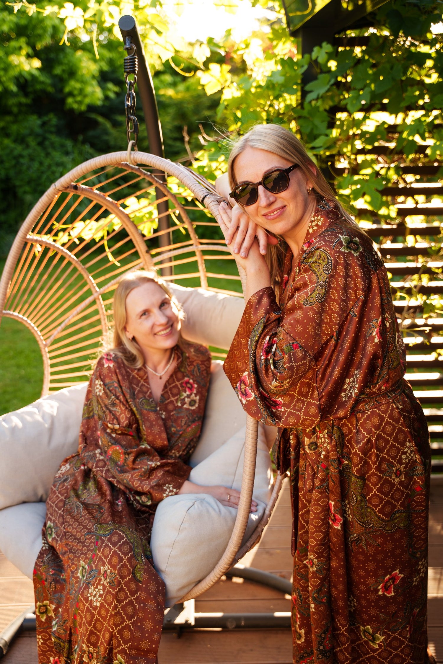 Two women - sisters wearing matching rust-brown Batik patterned kimono robes pose in a garden patio setting, with one sitting in a hanging wicker chair and the other standing beside it.