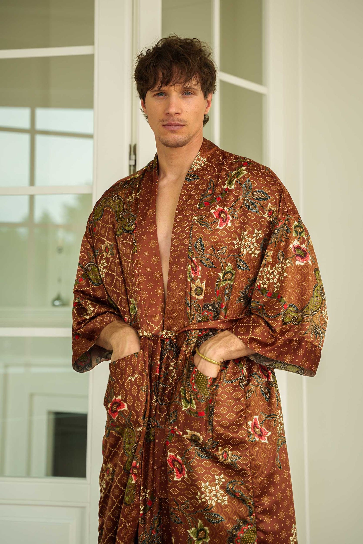Man wearing Toré Bumi kimono robe – long satin dressing gown in brown and olive batik pattern, hands in pockets.