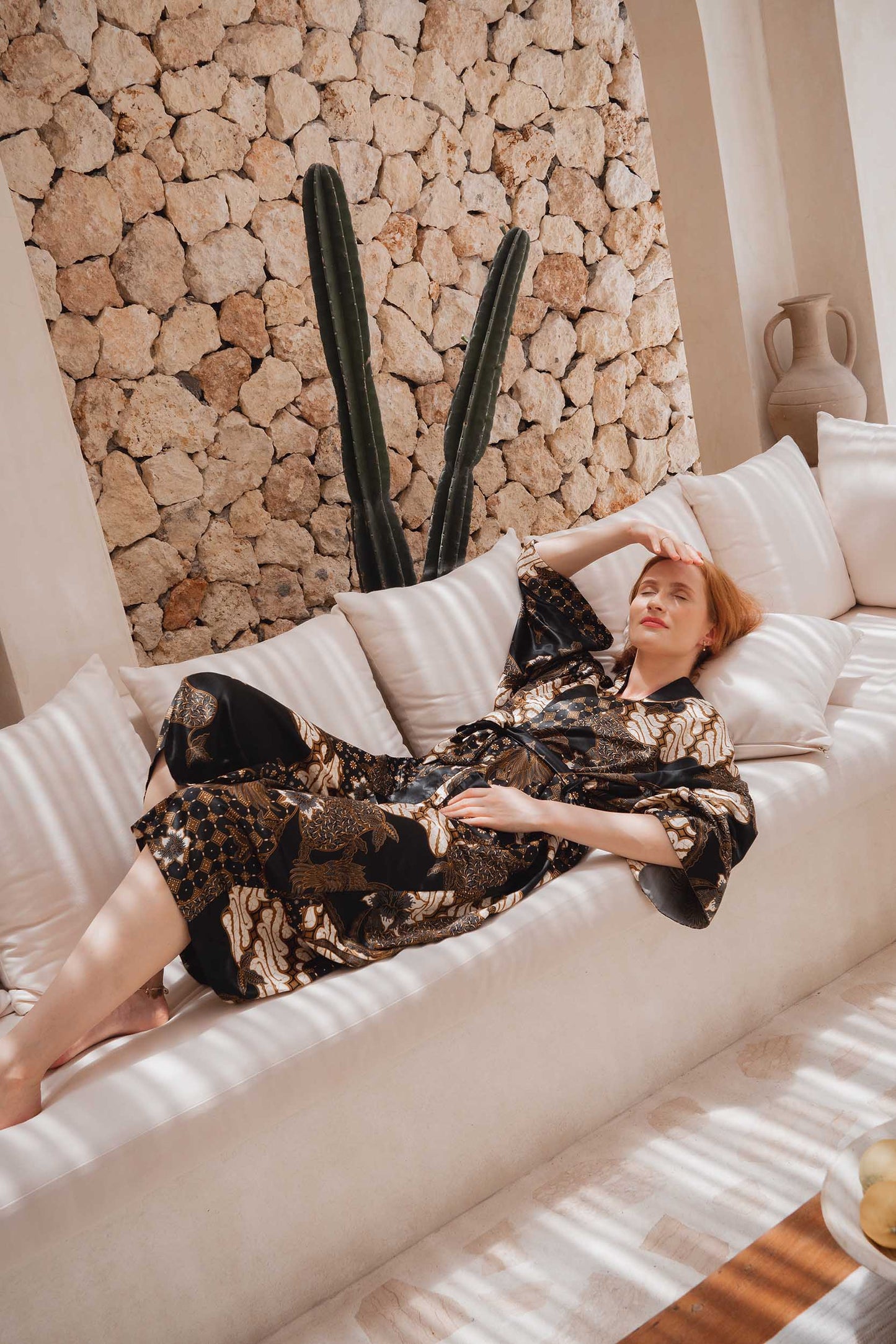 Woman wearing Toré Kencana robe – elegant long satin kimono for women in black and gold tones and laying on bed.