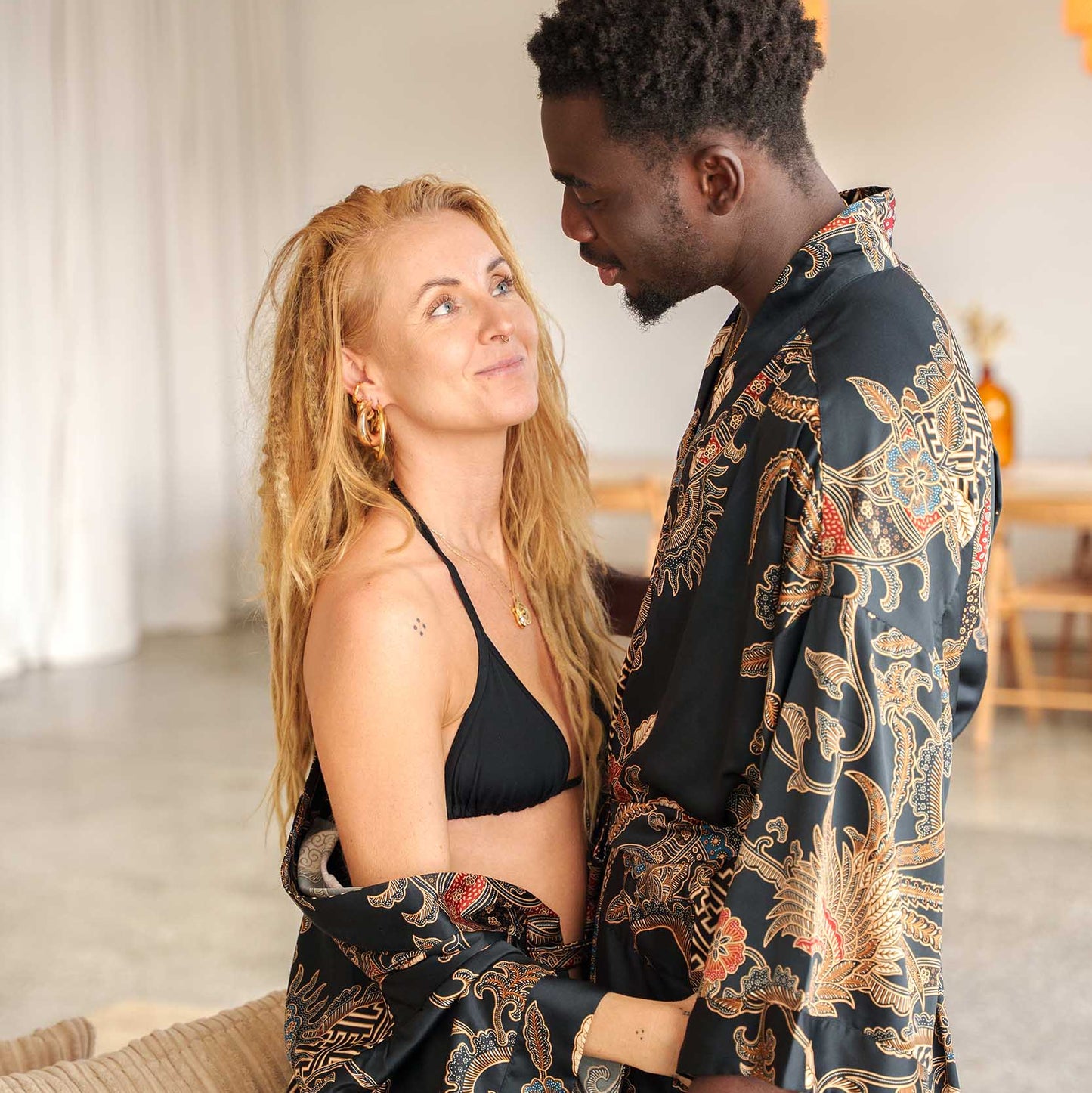 Couple wearing matching Toré Malam kimono robes – black satin dressing gowns with gold and red batik details.