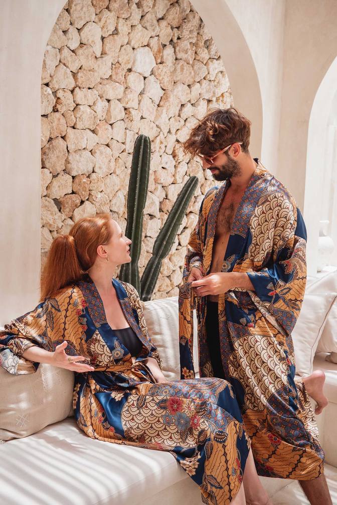 A man and a woman wearing matching patterned satin kimono robes in a luxurious room with a stone archway background. The robes feature a complex, ornate pattern of gold, blue, brown, and beige.