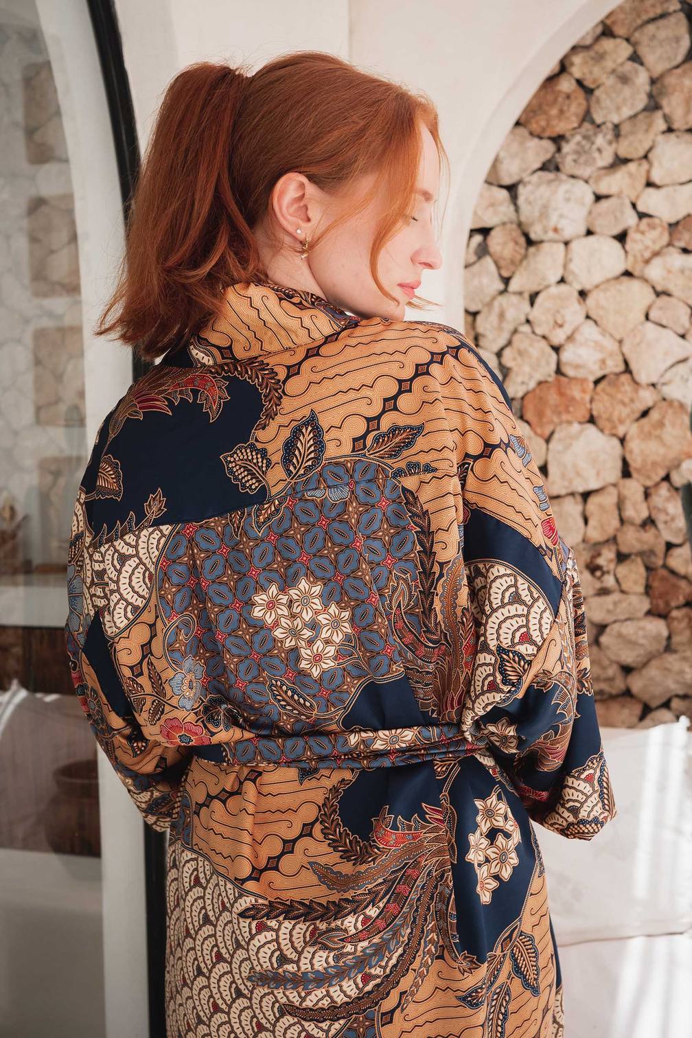 A woman with red hair tied back in a ponytail is shown from the back wearing a long-sleeve batik kimono robe with a tie waist. The robe features a complex multicolor pattern with navy blue, beige, brown, and red.