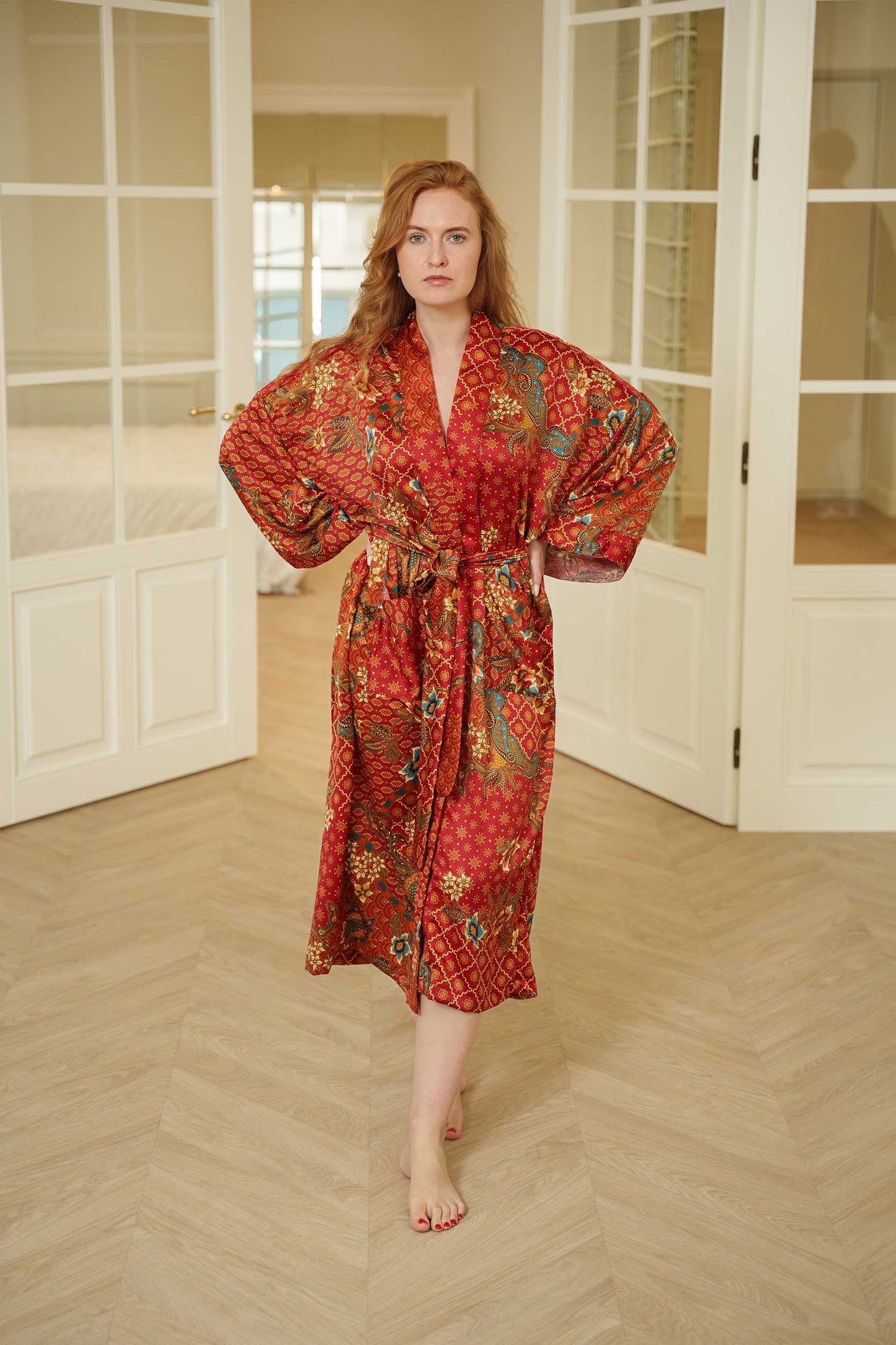 Woman wearing Toré Senja kimono robe – red satin long dressing gown with golden floral batik design.
