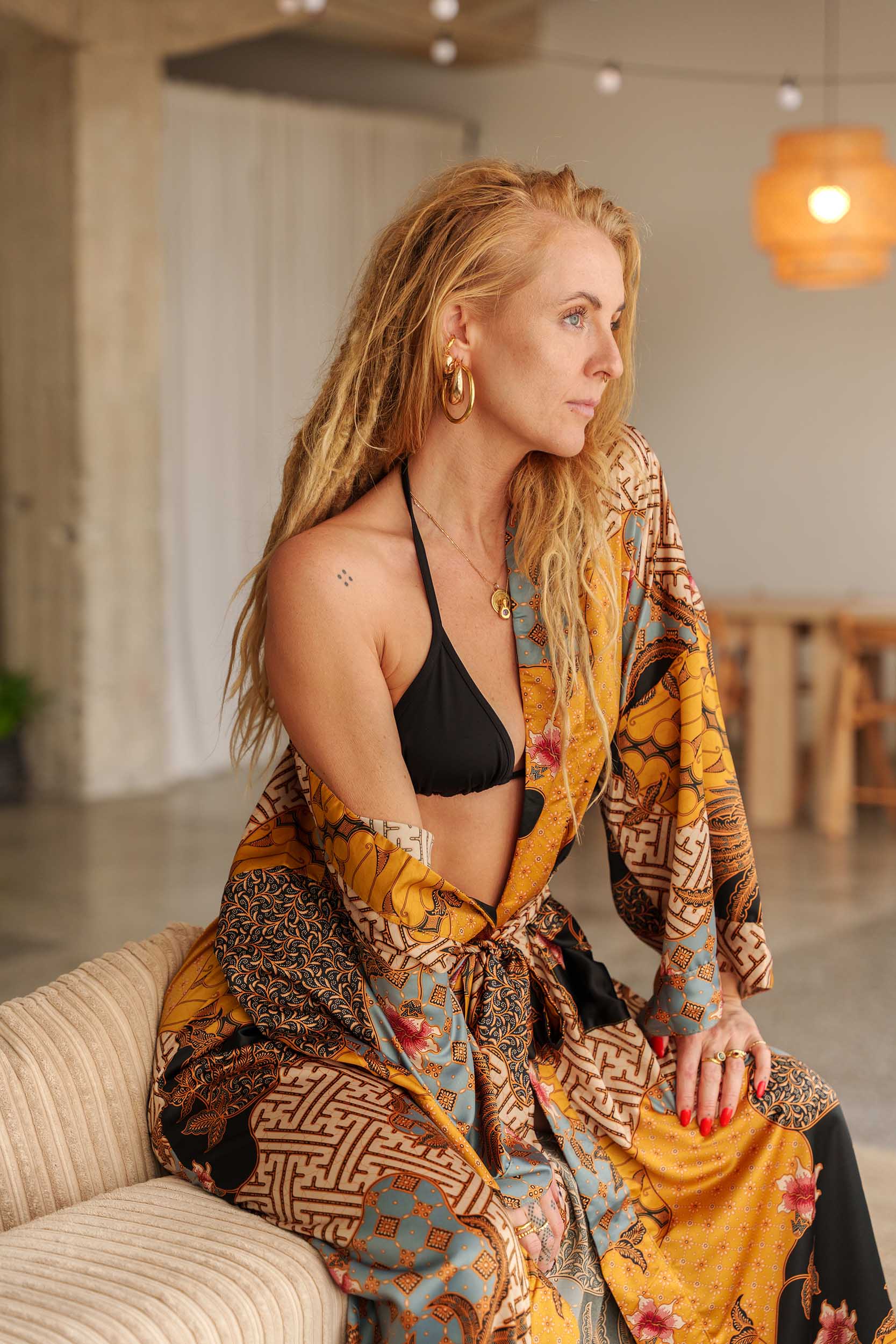 Woman wearing Toré Sorga kimono robe – long yellow and blue satin dressing gown with Indonesian batik design and sitting on a couch.