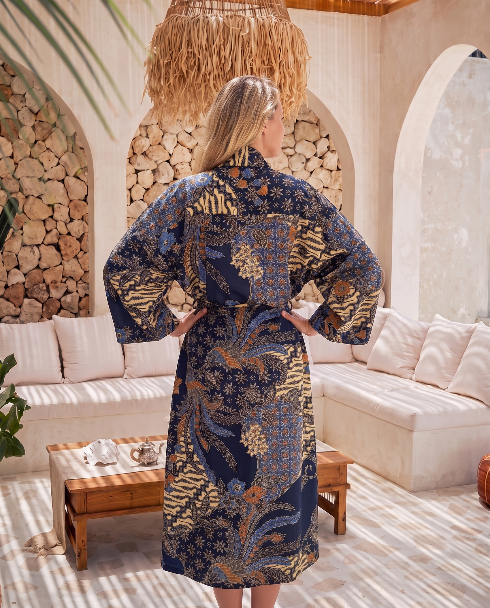 A woman stands with her back to the camera wearing a long-sleeved navy blue kimono robe with an intricate gold and beige floral pattern, set in a bright, modern bohemian-style lounge room.