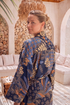 A woman wearing a navy blue satin kimono robe with a detailed orange, beige, and gold botanical Batik pattern, standing in a warm, sunlit lounge room with stone arches and white cushions.