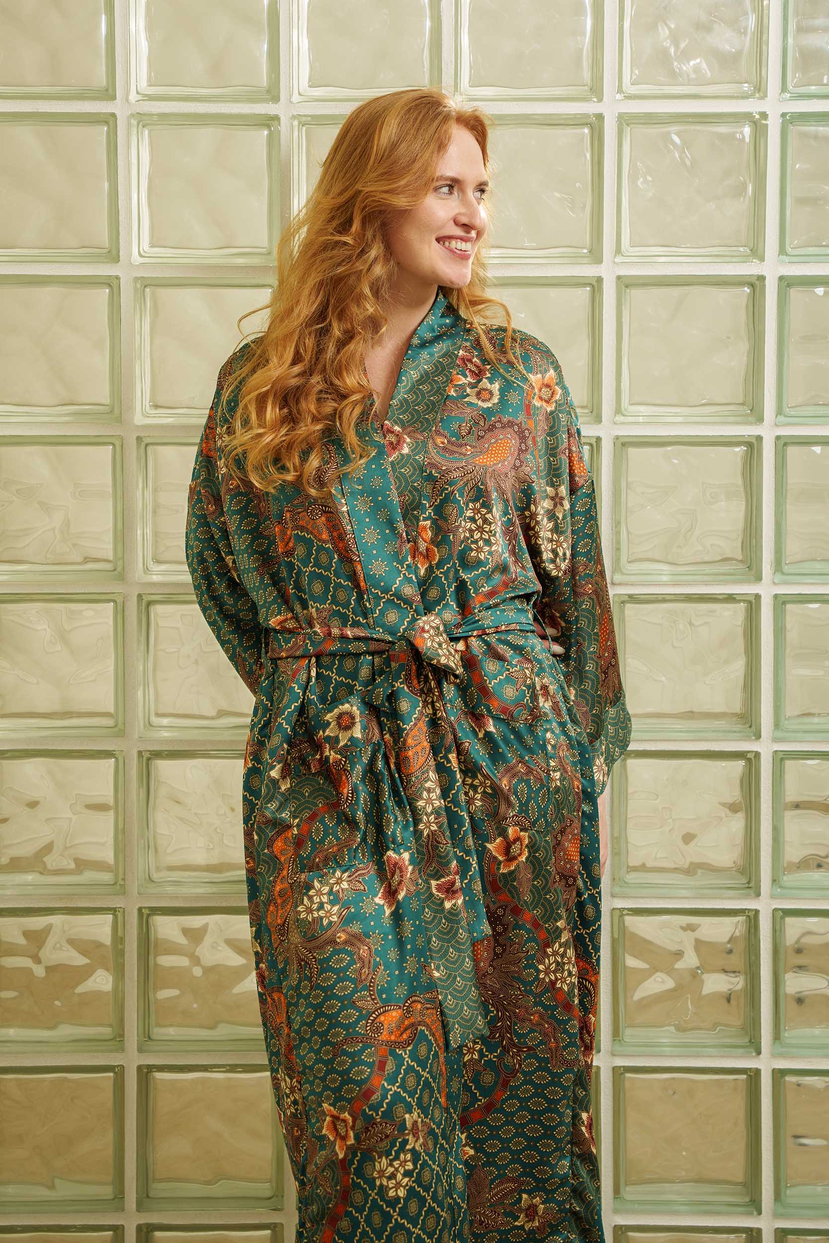 Woman wearing Toré Zamrud kimono robe – long emerald teal satin dressing gown with gold batik design.