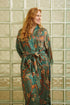 Woman wearing Toré Zamrud kimono robe – long emerald teal satin dressing gown with gold batik design.
